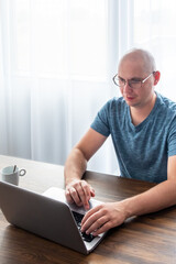Man in a blue t-shirt sitting at the table and using laptop. Remote work, work from home.