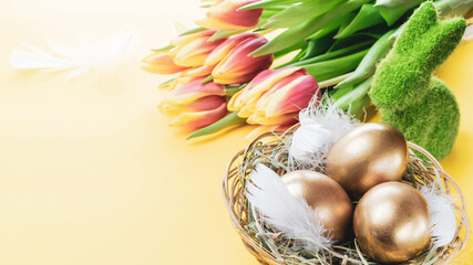 Golden easter colour eggs in basket with spring tulips, white feathers on pastel yellow background in Happy Easter decoration. Spring holiday concept.