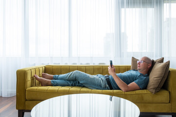 Man in a blue t-shirt lying on the yellow sofa and using smartphone. Remote work, work from home.
