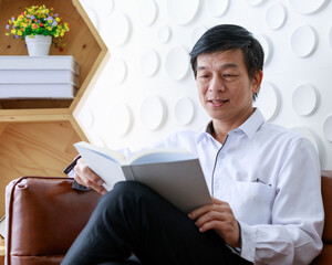 Asian old middle aged confident black and gray hair male businessman wears white long sleeve shirt black pants glasses in pocket sit on brown leather couch having problem to read white book in hands