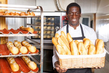 Portrait of successful baker holding wicker basket with fresh baguettes in kitchen of his bakery