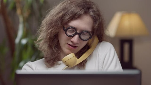 Portrait of intelligent concentrated 1980s man talking on yellow vintage telephone sitting with retro laptop indoors. Genius Caucasian young man at home or in office.