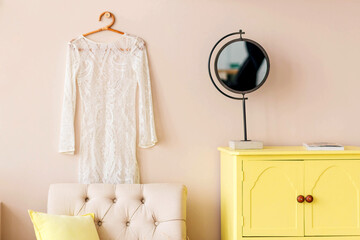 A yellow chest of drawers, an armchair, a mirror stand against a background of a beige wall.