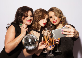 Three fashionable young women dressed in black cocktail dresses are singing with a microphone, holding disco balls, glasses of champagne and smiling