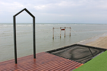 Door frame, wooden decking, and net seat overhang a beach with a swing in the shallows of the ocean; activities for tourists.