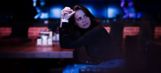Beautiful sad serious glamorous young woman sitting in a night cafe alone