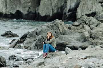 young woman in jeans and red sneakers sits on rocks near the stormy oceans with waves. local tourism, solo travel. mental health communication with nature.