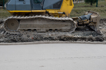 Crawler bulldozer with yellow cab with exposed blade and mud-spattered tracks stands on excavated soil surface of ground next to strip of path rolled up by road roller at road works site.