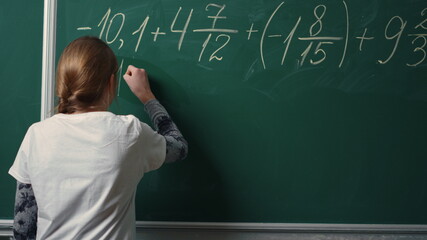 Back view of school girl doing math task on blackboard in class.Student writing 