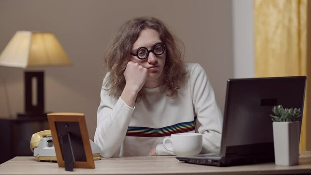 Frustrated depressed young man in retro eyeglasses sitting at table with vintage laptop and telephone looking at picture and thinking. Portrait of sad Caucasian nerd guy indoors at home.