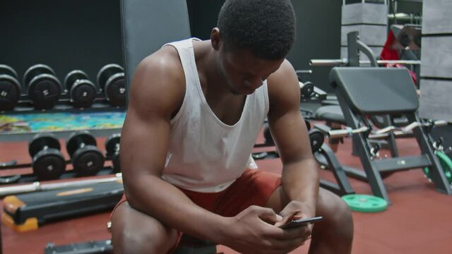 Young muscular afro man sits on bench, uses phone and relaxes after exercise