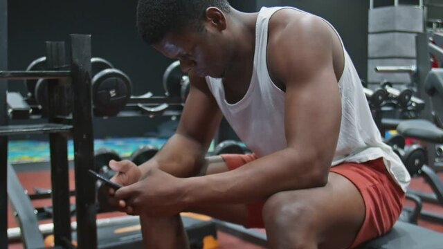 Side view of young muscular afro man sits on bench, uses phone and relaxes after exercise