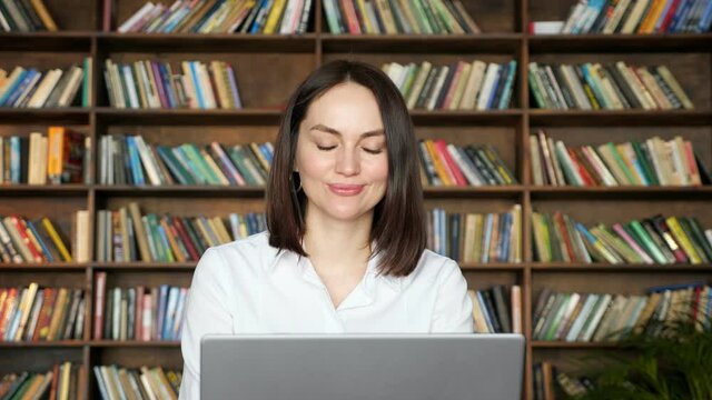 Woman freelancer with short hair talks online on near grey laptop against large bookshelves in local librar
