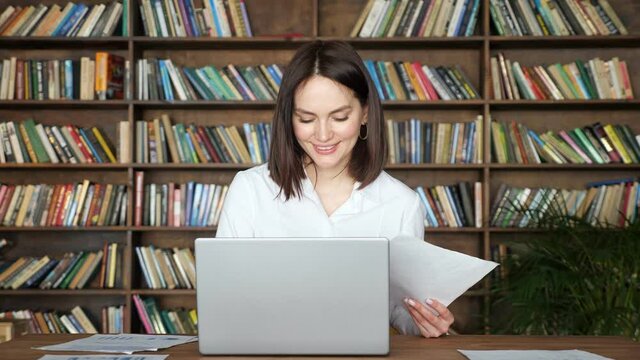 Smiling young woman in stylish white blouse works with papers near contemporary laptop at table against large library racks full of book