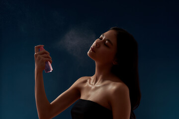 Young attractive asian woman spraying thermal water on her face while standing against dark blue...