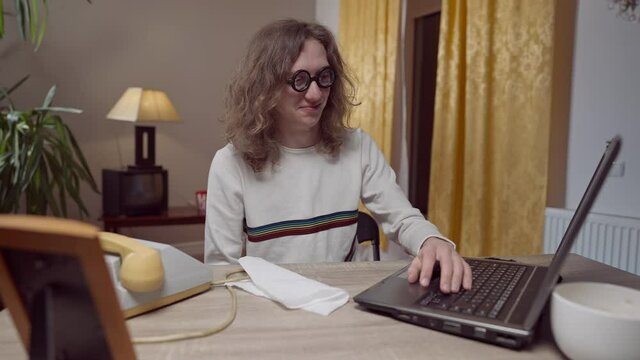 Young nerd 1980s 1990s man using vintage outdated laptop moving hand under table. Middle shot portrait of Caucasian geek with long curly hair resting at home indoors. Retro lifestyle.