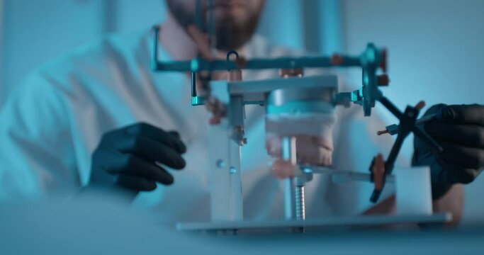 Close-up an axiosplit device inside of it is standing a ceramic model of an upper jaw and male medical worker wearing at black latex gloves and disposable eyeglasses is putting on the device a detail.