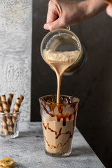 Hand holding jar and pouring milkshake drink into the glass decorated with caramel.