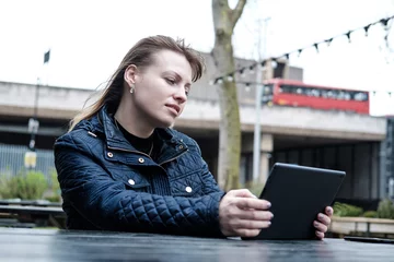 Foto op Plexiglas Jonge vrouw met haar tablet zittend aan tafel op een terras in de buurt van Waterloo bridge in Londen. Smartphone ligt ook op tafel. Een rode bus wordt weerspiegeld op het glas. © Jorge Elizaquibel