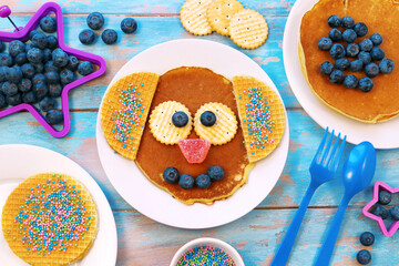 Funny pancakes for children. Pancake in a shape of dog on white plate, blue wooden background, top view. Creative idea for kids breakfast.	