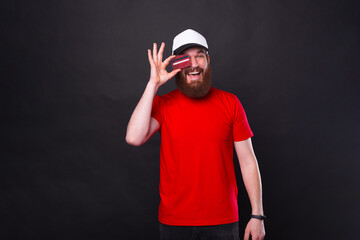 Naklejka premium Cheerful young bearded hipster man wearing red t-shirt and holding red credit card over eyes