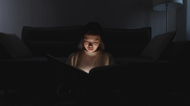 Night time, portrait of asian female sitting at home on the floor and reading a book, warm light, self-isolation during the pandemic, mystical lighting effect.