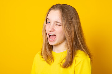 Childish and playful teenage girl winking at camera, keeping mouth wide opened on yellow background...