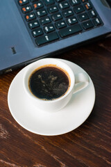 Coffee in a white cup and a laptop on the background on a brown wooden table. Concept of remote work or training, the start of the working day in the office.