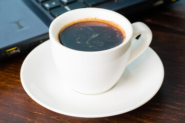 Coffee in a white cup and a laptop on the background on a brown wooden table. Concept of remote work or training, the start of the working day in the office.