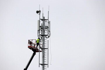 Worker repairs cell tower standing on lifting platform. Mobile telecommunication equipments on sky...