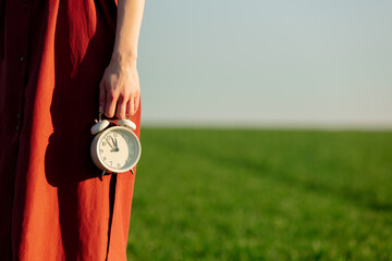 Woman in red dress hold vintage alarm clock on green wheat field in background