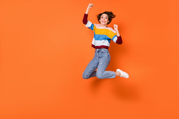 Full length photo of delighted girl fists up celebrate jumping high isolated on orange color background