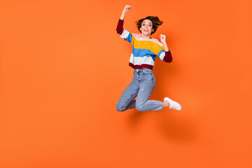 Full length photo of delighted girl fists up celebrate jumping high isolated on orange color...