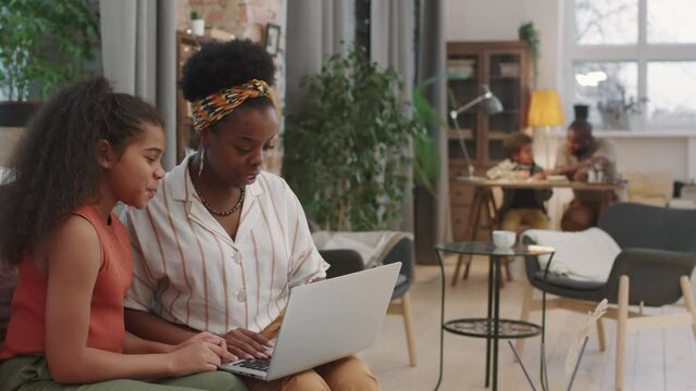 Panning medium shot of african-american woman helping elder daughter with online studies using laptop together sitting on sofa while father and little son playing in background in cozy living room
