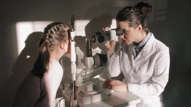 Zoom-in medium shot of professional young female optometrist having little girl eyesight checked sitting at table with phoropter machine at sunlight filled doctor office