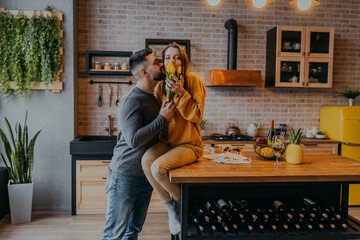 Bearded man presents flowers to woman in the kitchen. Woman sitting on table. Man kisses woman in kitchen.