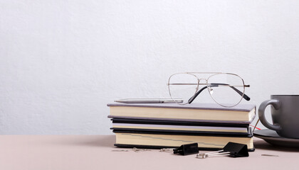 Closeup of stack of notebooks, eye glasses, cup of coffee, stationary binder clips on the desk against white empty wall