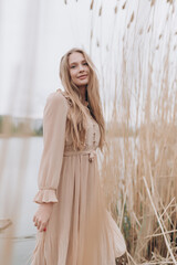 a beautiful girl with brown hair in a beige dress stands in the tall reeds by the water
