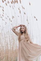 a beautiful girl with brown hair in a beige dress stands in the tall reeds by the water