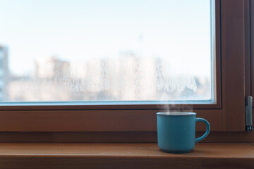 Blue mug with hot drink standing on windowsill by the window inside the room in the morning. Copy space. Concept of an invigorating drink, good morning.