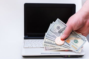 Hand holds Bitcoin and one hundred dollar paper bills over laptop with black screen on white background.
