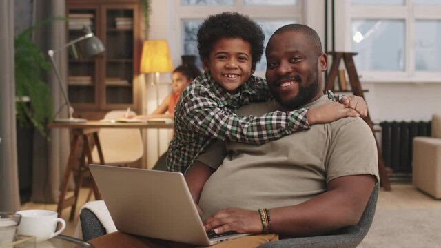 Slow-motion medium portrait of loving afro-american dad and little son looking at each other hugging then smiling to camera spending evening together at home. Elder sister doing homework in background