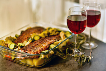 two glasses of red wine and a large glass dish with baked fish and vegetables