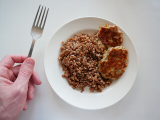 Buckwheat with a chicken cutlet on a plate and a hand with a fork on the left. White background 