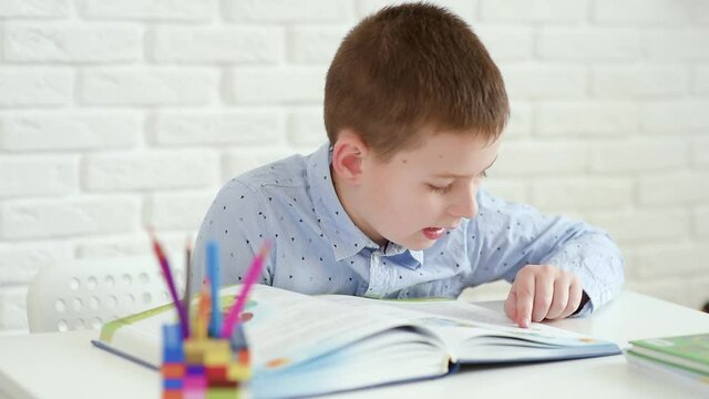 schoolboy child reads a book sitting at a white table on a white brick background