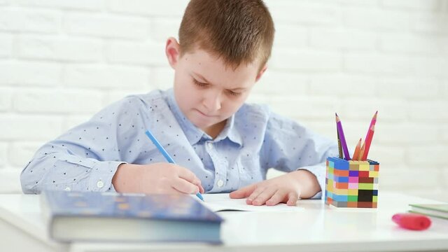 schoolboy writes in a notebook sitting at a white table.