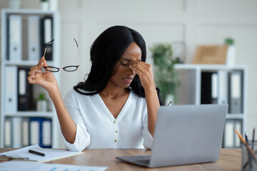 Black young businesswoman having tired irritated eyes, sitting at desk with laptop, exhausted from...