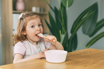 A little girl eats porridge from a white bowl, a girl has breakfast sitting at the table. Healthy breakfast, healthy food