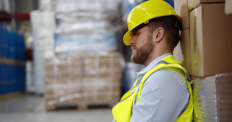 Exhausted young cargo worker sleeping leaning on large container in warehouse