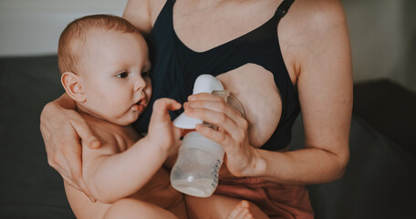 Young mother with newborn cute infant naked baby boy, holding him on arms, using breast pump hugging and breastfeeds with breast milk. Healthy child, concept of hospital and happy motherhood. Nursery.